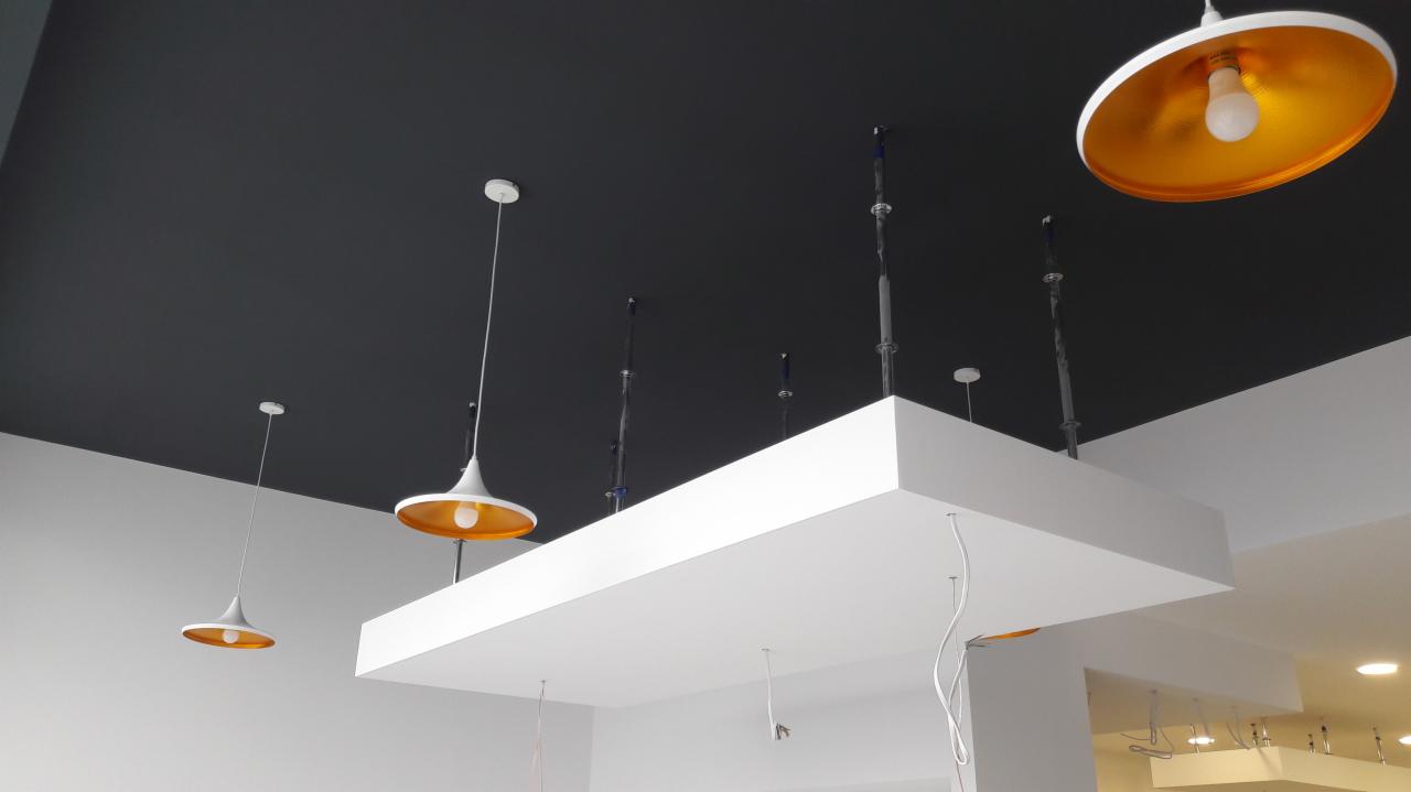 Ciemnoszary sufit z zamontowanymi białymi lampami wiszącymi z pomarańczowym wnętrzem klosza i białą podwieszaną konstrukcją z widocznymi przewodami elektrycznymi w trakcie instalacji.