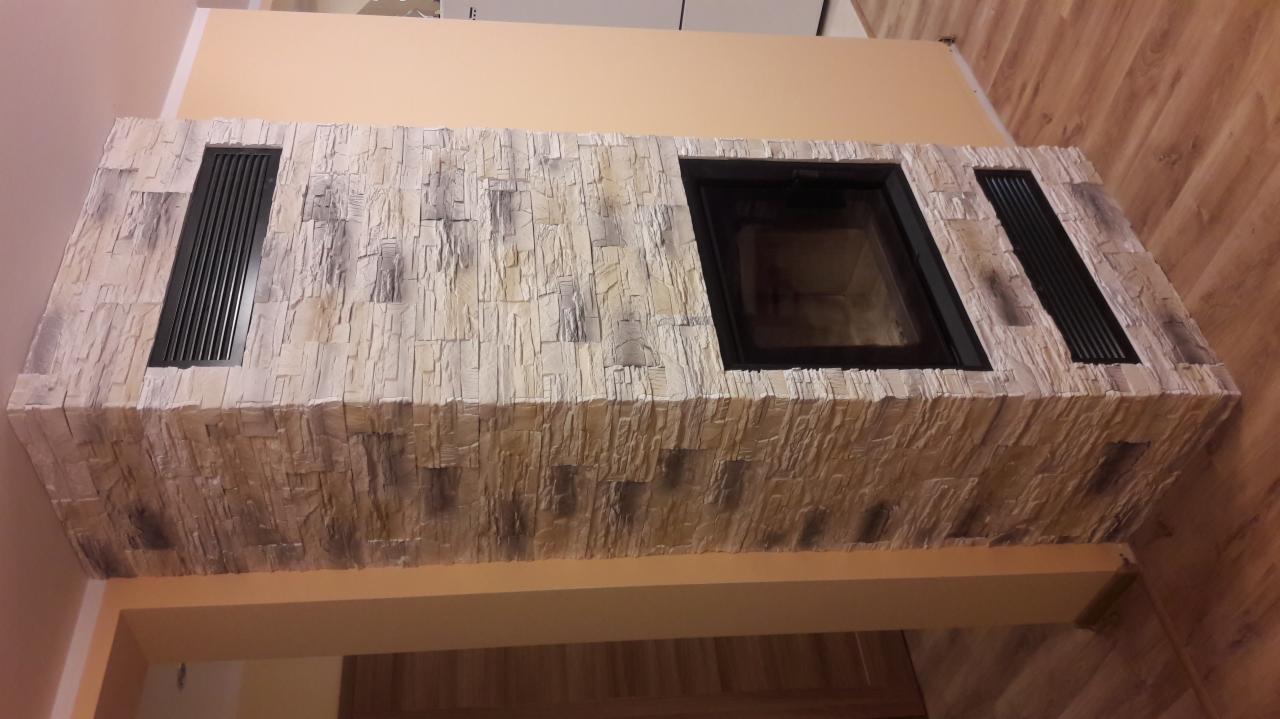 Obudowa kominka wykończona kamieniem dekoracyjnym w odcieniach beżu i szarości, z czarnym wkładem kominkowym i kratkami wentylacyjnymi, na tle beżowej ściany i drewnianej podłogi.