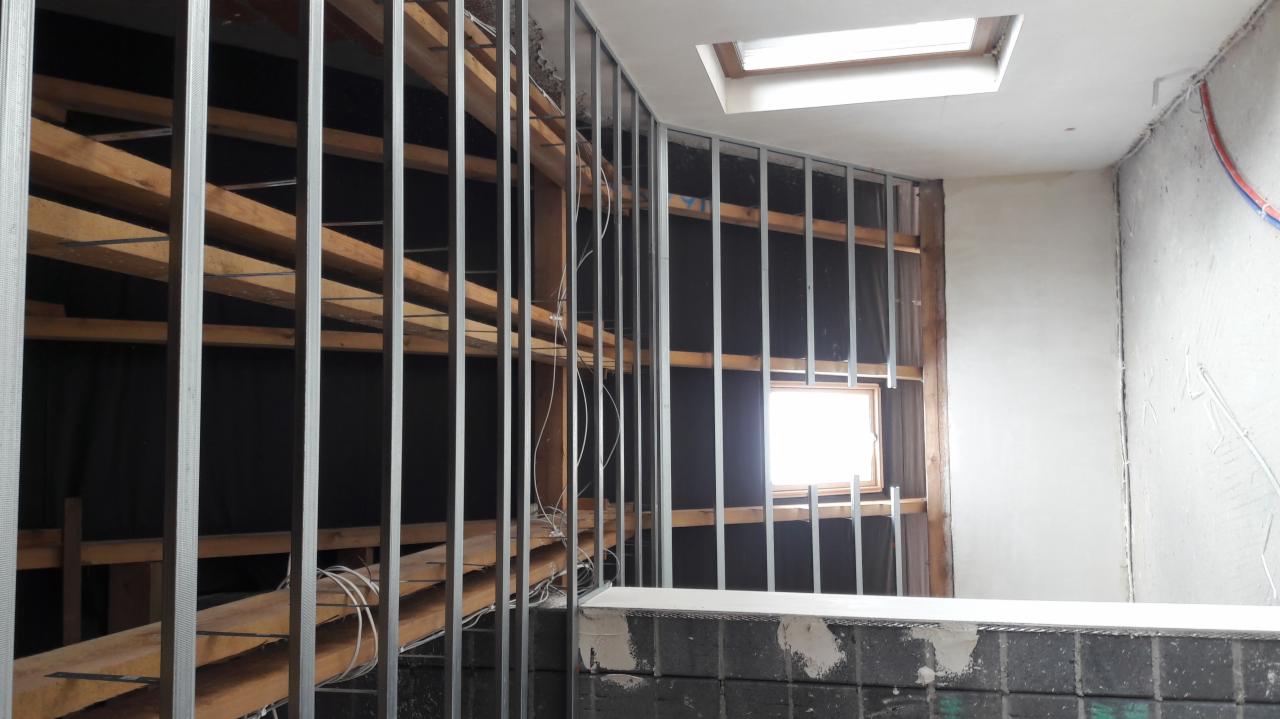 Wnętrze w trakcie prac: metalowa konstrukcja pod płytę gipsową na drewnianym ruszcie przy oknie, fragment otynkowanej ściany z instalacją elektryczną i okno dachowe.