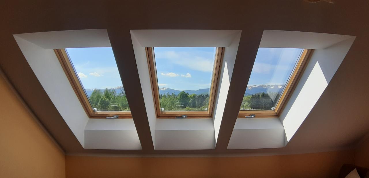 Trzy okna dachowe z drewnianymi ramami, przez które widać błękitne niebo, zielone drzewa i odległe góry.