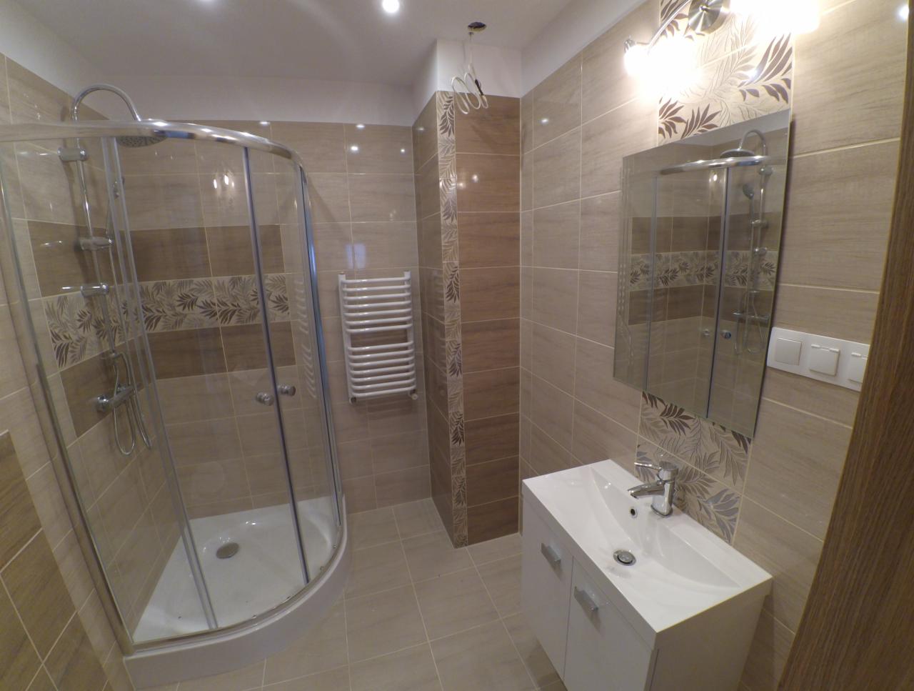 Wykończona łazienka w beżowych odcieniach z kabiną prysznicową, grzejnikiem drabinkowym i umywalką z szafką, z dekoracyjnym pasem płytek z motywem liści.