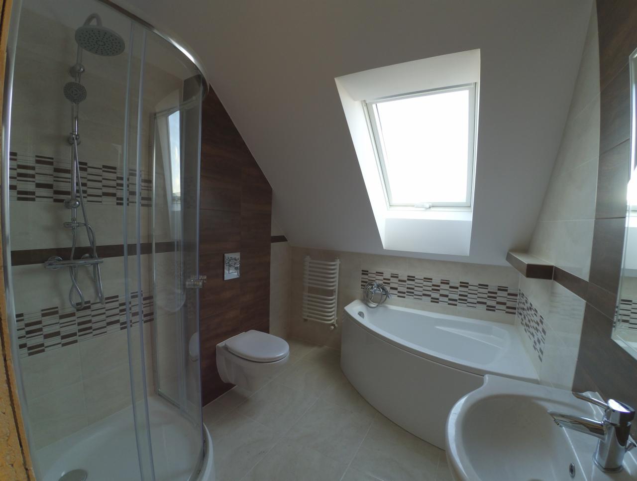 Jasna łazienka na poddaszu z kabiną prysznicową, asymetryczną wanną i oknem dachowym, wykończona beżowymi i brązowymi płytkami z dekoracyjnym pasem mozaiki.