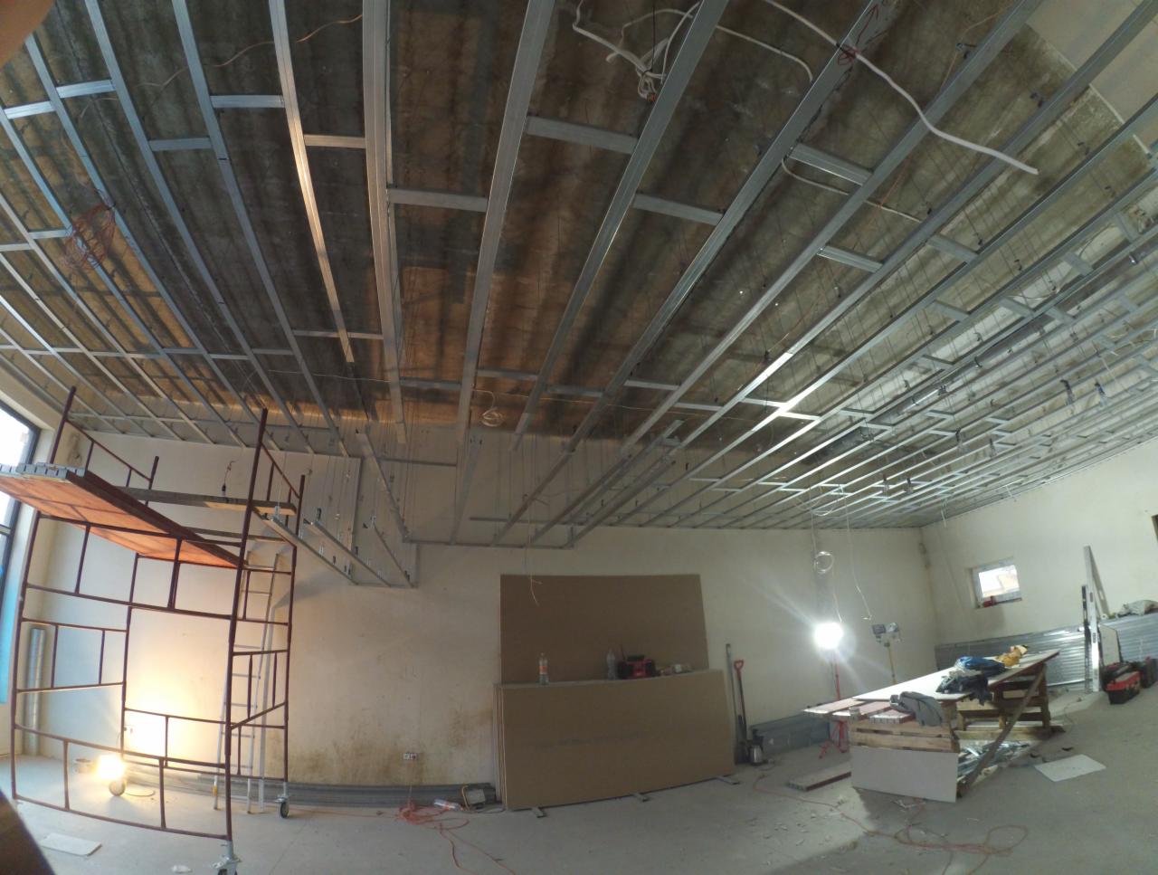 Stelaż sufitu podwieszanego z profili metalowych, widok z dołu, widoczne kable i płyty kartonowo-gipsowe w tle, rusztowanie z lewej strony.