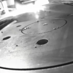 Metalowy element z wywierconymi otworami i śladami obróbki, widoczne drobne opiłki i zarysowania, wykonany w odcieniach szarości, z ostrym światłem odbijającym się od powierzchni.