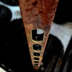 Zardzewiała, perforowana listwa stalowa ustawiona pionowo na tle ciemnych, okrągłych elementów maszynowych.