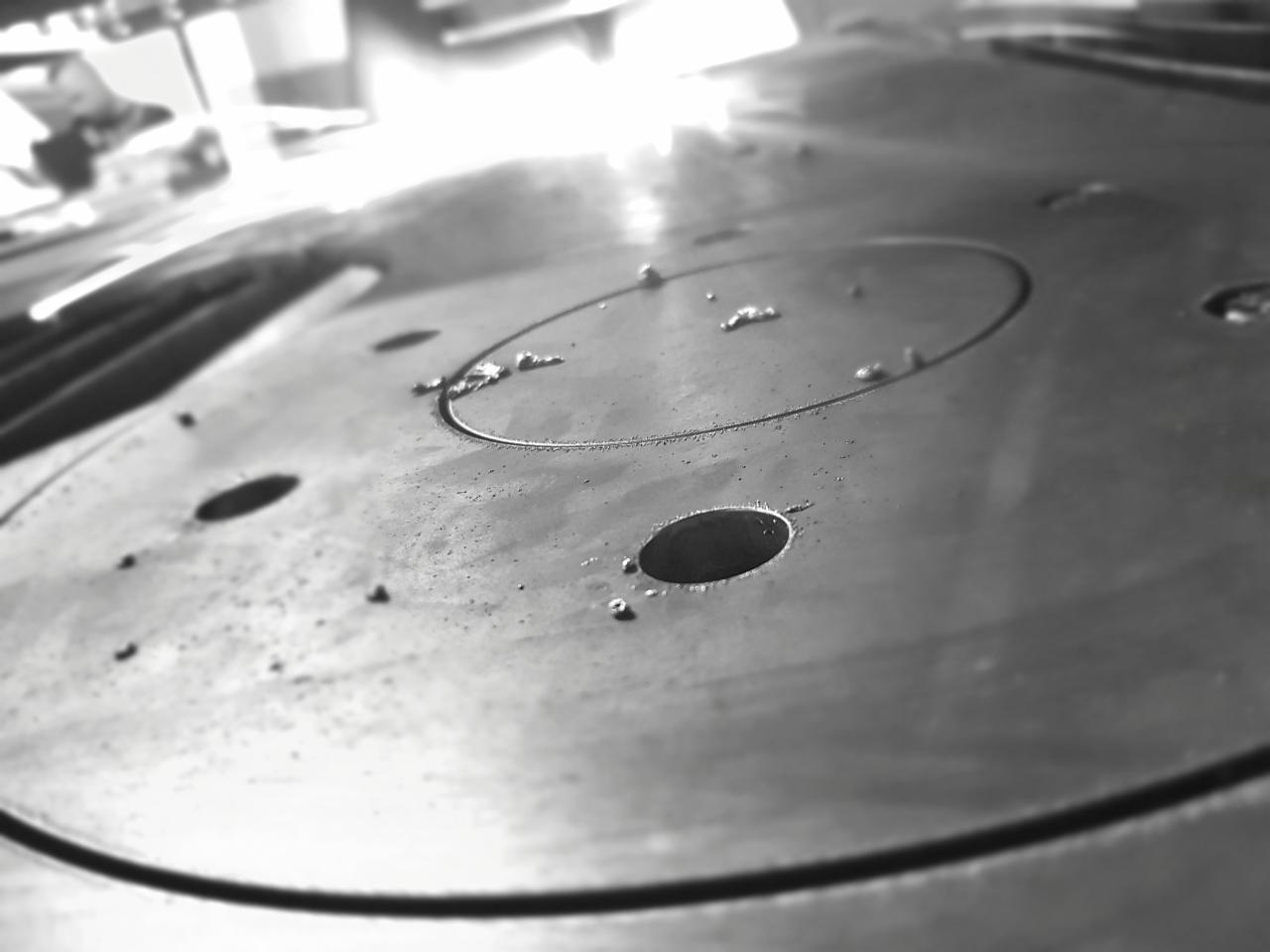 Metalowy element z wywierconymi otworami i śladami obróbki, widoczne drobne opiłki i zarysowania, wykonany w odcieniach szarości, z ostrym światłem odbijającym się od powierzchni.