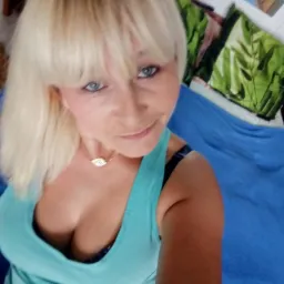 Kobieta z blond włosami w błękitnej bluzce na ramiączkach, uśmiechnięta, z delikatnym makijażem i złotym naszyjnikiem. Ujęcie od dołu.