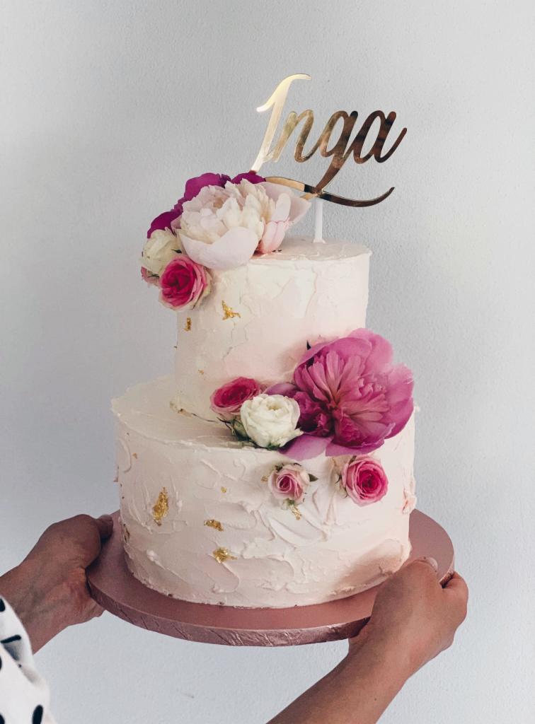 Dwupoziomowy tort weselny w odcieniach bieli i różu, zdobiony świeżymi piwoniami i różami oraz złotymi płatkami, z topperem z imieniem 'Inga', trzymany przez dwie osoby.