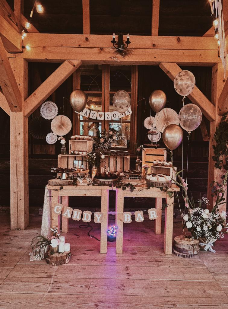 Rustykalny stół z napisem 'Candy Bar' udekorowany balonami, kwiatami i słodkościami w drewnianej chacie, przygotowany na przyjęcie.
