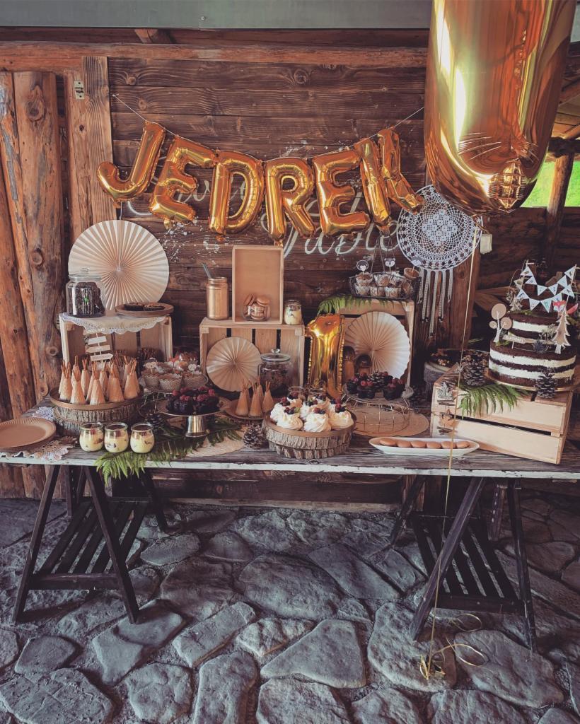 Rustykalny stół z deserami i dekoracjami urodzinowymi, w tym balonami z napisem 'JĘDREK', tortem warstwowym i różnorodnymi słodkościami na drewnianych podstawkach i w szklanych naczyniach, całość...
