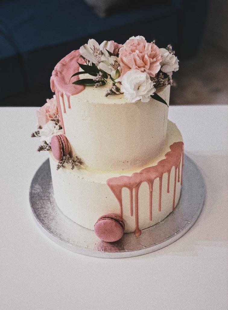 Dwupoziomowy tort z białym kremem, różowym polewą i dekoracją z żywych kwiatów oraz makaroników, umieszczony na srebrnej tacy.