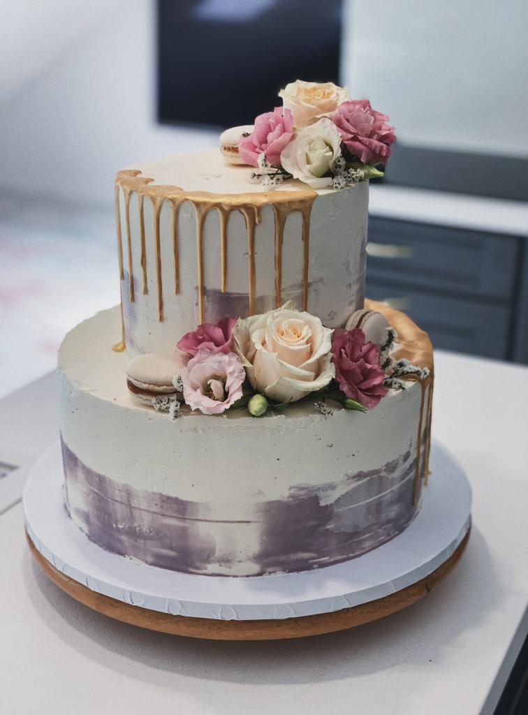 Dwupoziomowy tort weselny z białym kremem, złotymi zaciekami, dekorowany różowymi i białymi różami oraz makaronikami, umieszczony na drewnianej podstawie.