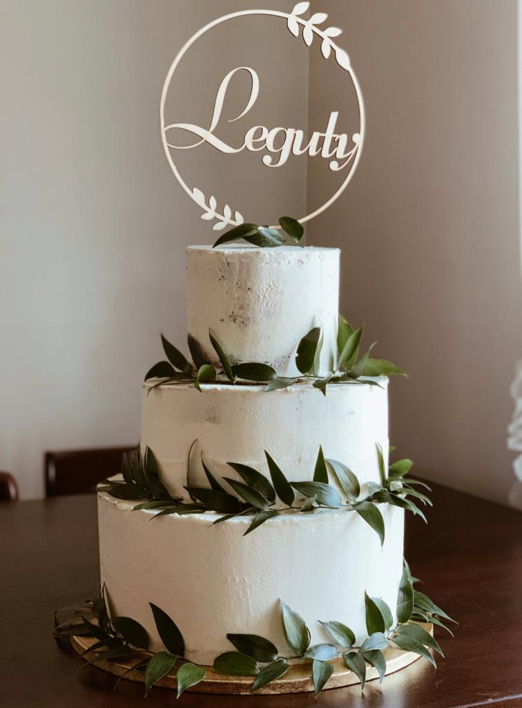 Trzypiętrowy tort weselny z białym kremem, ozdobiony świeżymi zielonymi liśćmi i drewnianym topperem z napisem 'Leguty' w okrągłej ramce z motywem liści.