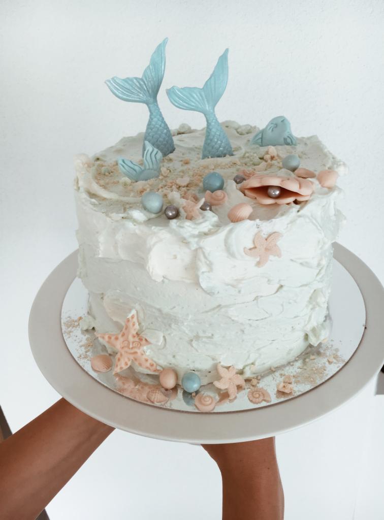Tort zdobiony motywem morskim: dwie błękitne płetwy syreny, muszelki z perłami, rozgwiazdy i posypka imitująca piasek na białym kremie, trzymany na srebrnej tacy.