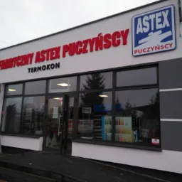 Fasada budynku z dużymi oknami, nad wejściem czerwony napis 'Punkt Fabryczny Astex Puczyński Termokon' i logo Astex Puczyński w niebieskiej ramce.