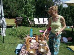 Kobieta w okularach przygotowuje zestaw mis tybetańskich na wzorzystym obrusie podczas sesji relaksacyjnej na trawie w ogrodzie, w tle leżanka i osoba odpoczywająca.