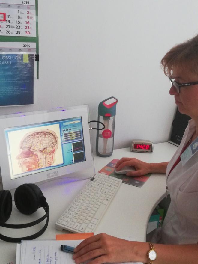 Kobieta w okularach pracuje przy biurku z komputerem wyświetlającym przekrój głowy i mózgu, obok butelka z wodą i notatnik z długopisem