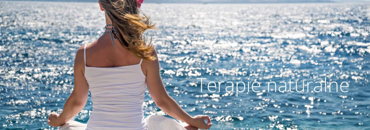 Kobieta siedząca w pozycji lotosu na tle morza, z widocznymi promieniami słońca odbijającymi się od wody; włosy rozwiane na wietrze, na głowie kwiat, na szyi sznur koralików.