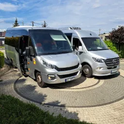 Dwa srebrne busy: Iveco z otwartymi drzwiami i Mercedes, stojące na ozdobnym brukowanym podjeździe w słoneczny dzień.