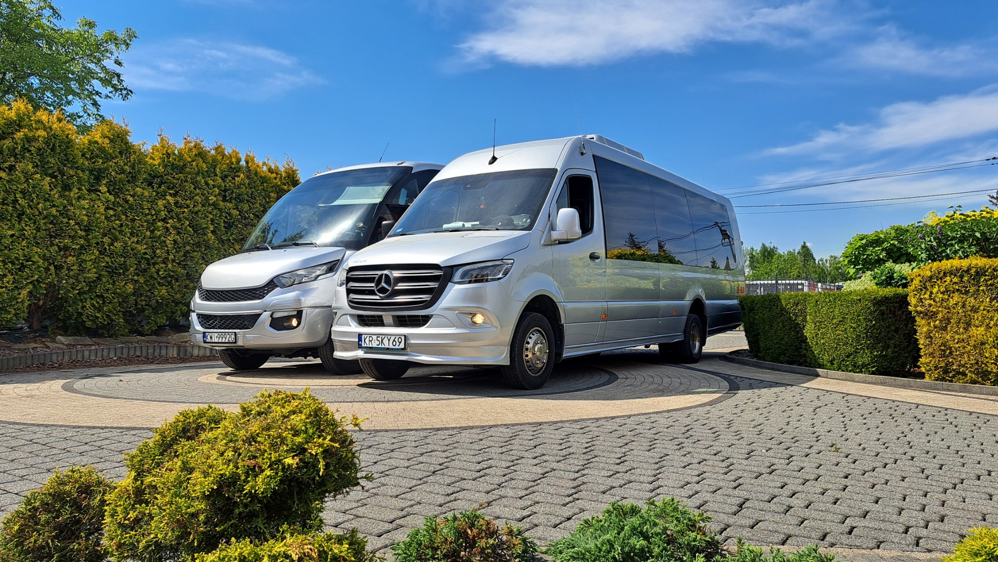 Dwa srebrne busy, jeden marki Iveco z widoczną tablicą rejestracyjną KWI 9992G, drugi Mercedes-Benz z tablicą KR 5KY69, zaparkowane na kostce brukowej przed zielonymi tujami w słoneczny dzień.