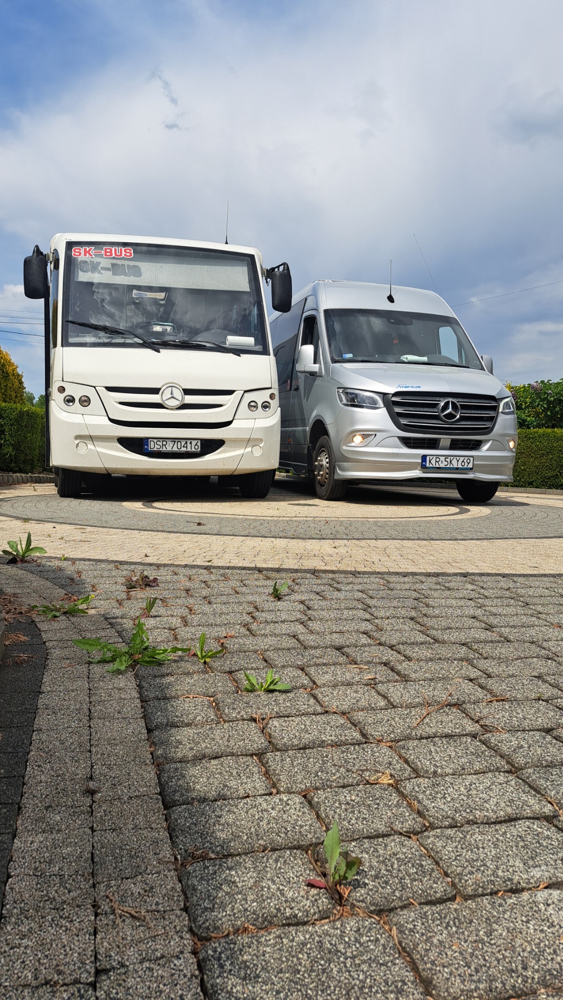 Dwa busy marki Mercedes-Benz: biały SK-BUS i srebrny, stojące obok siebie na kostce brukowej z chwastami, na tle zieleni i błękitnego nieba.