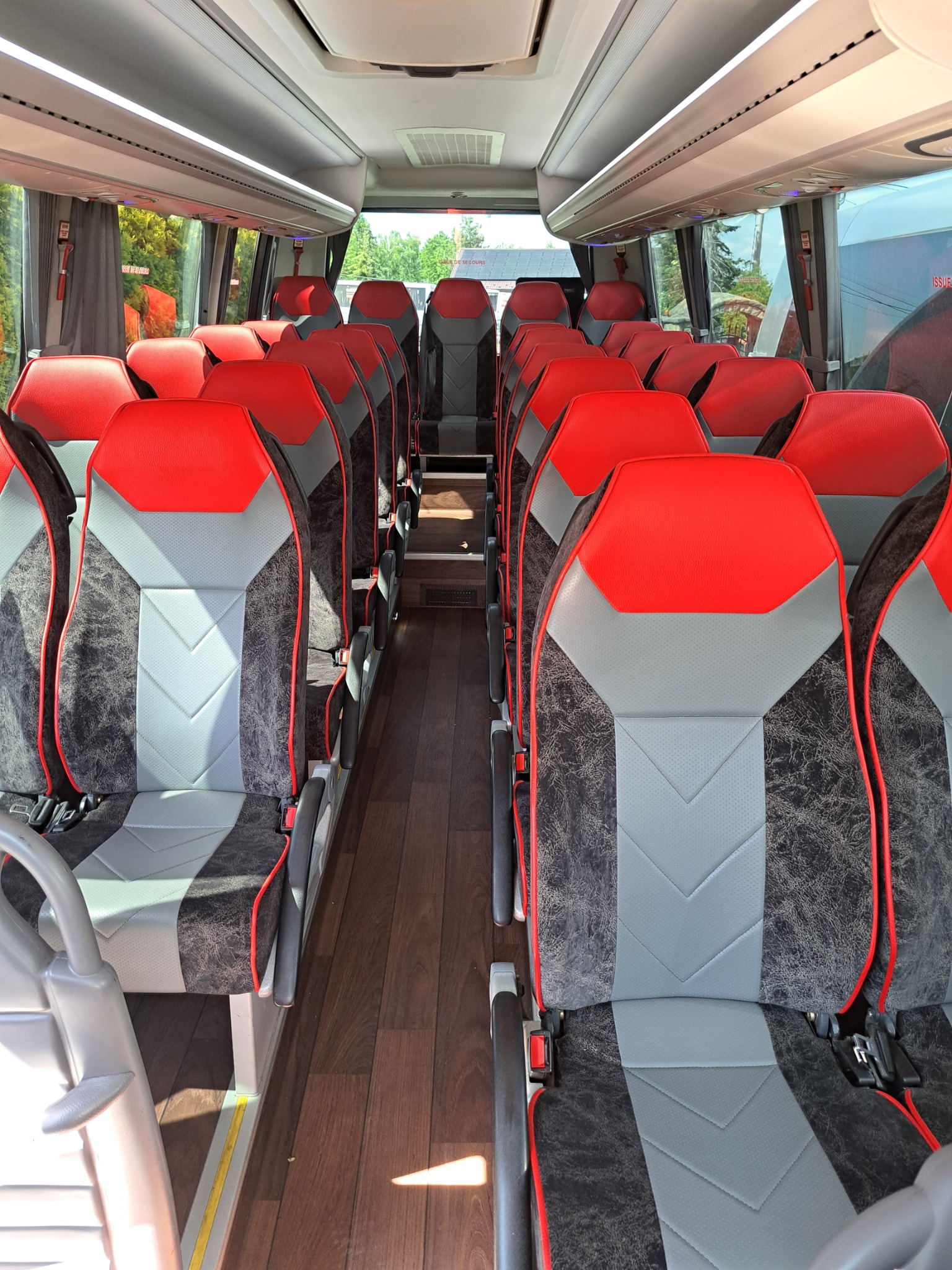 Wnętrze autobusu turystycznego z rzędami foteli w kolorze czerwonym i szarym, widok z przodu na przejście i drewnianą podłogę.
