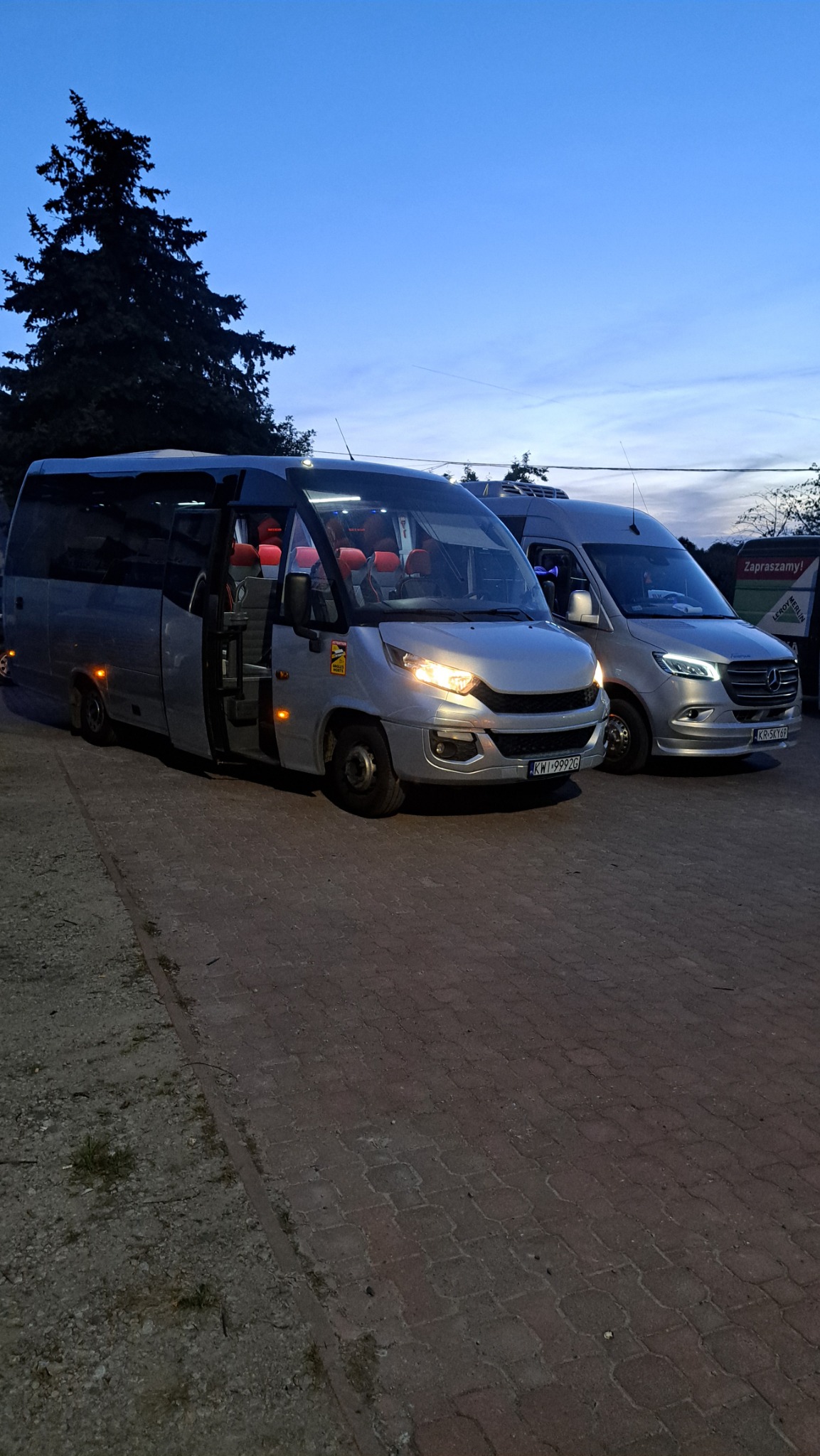 Dwa srebrne busy zaparkowane obok siebie na brukowanej powierzchni o zmierzchu, z otwartymi drzwiami w jednym z nich, ukazując czerwone fotele wewnątrz.