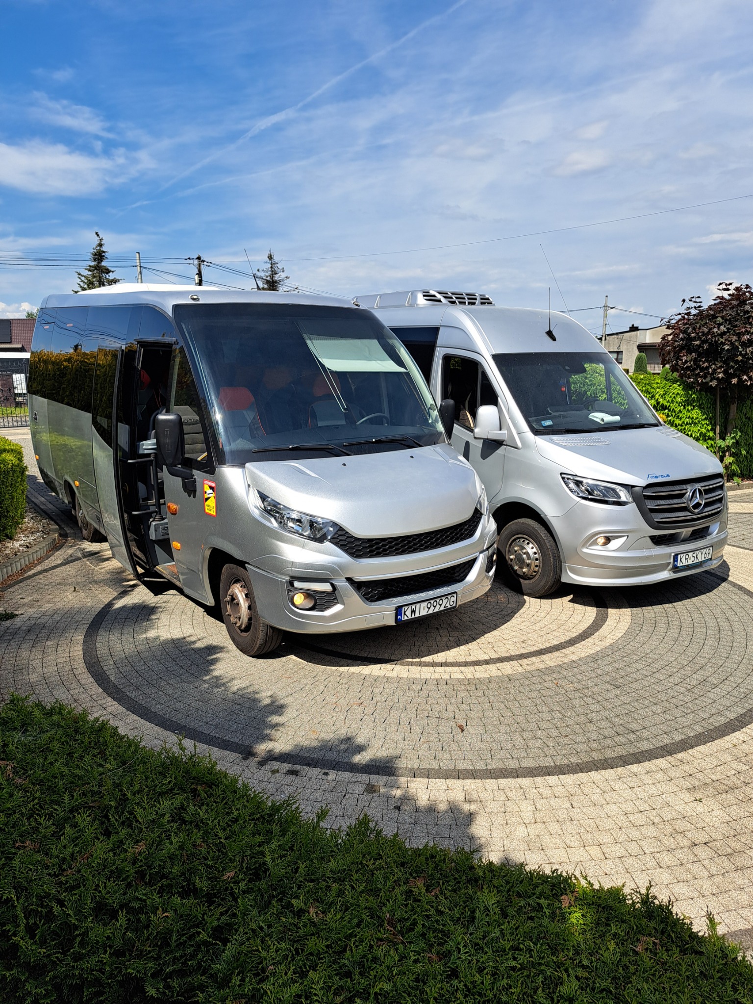 Dwa srebrne busy: Iveco z otwartymi drzwiami i Mercedes, stojące na ozdobnym brukowanym podjeździe w słoneczny dzień.