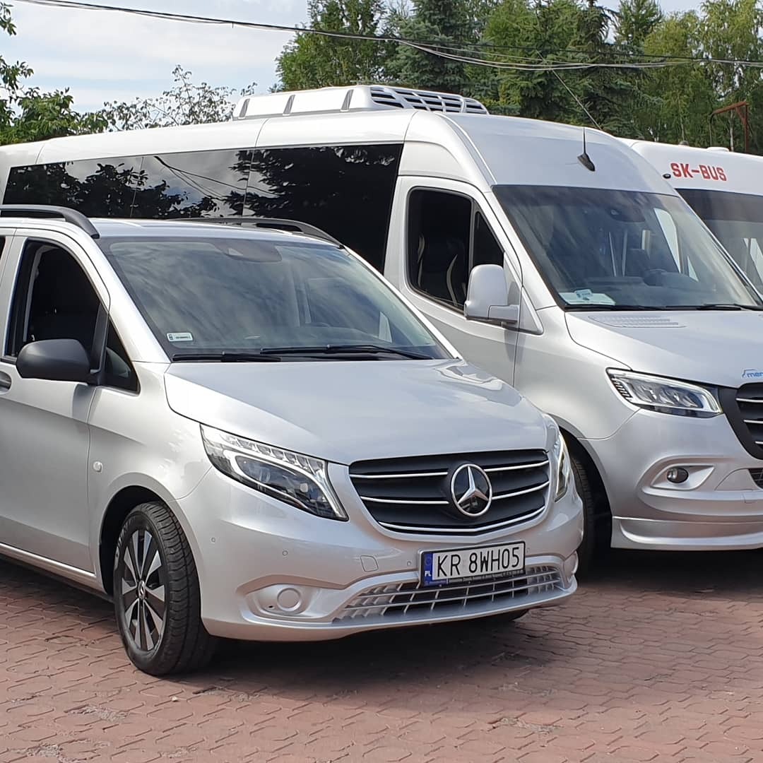 Dwa srebrne busy Mercedes-Benz, jeden mniejszy z relingami dachowymi i drugi większy z napisem SK-BUS, stojące na czerwonej kostce brukowej.