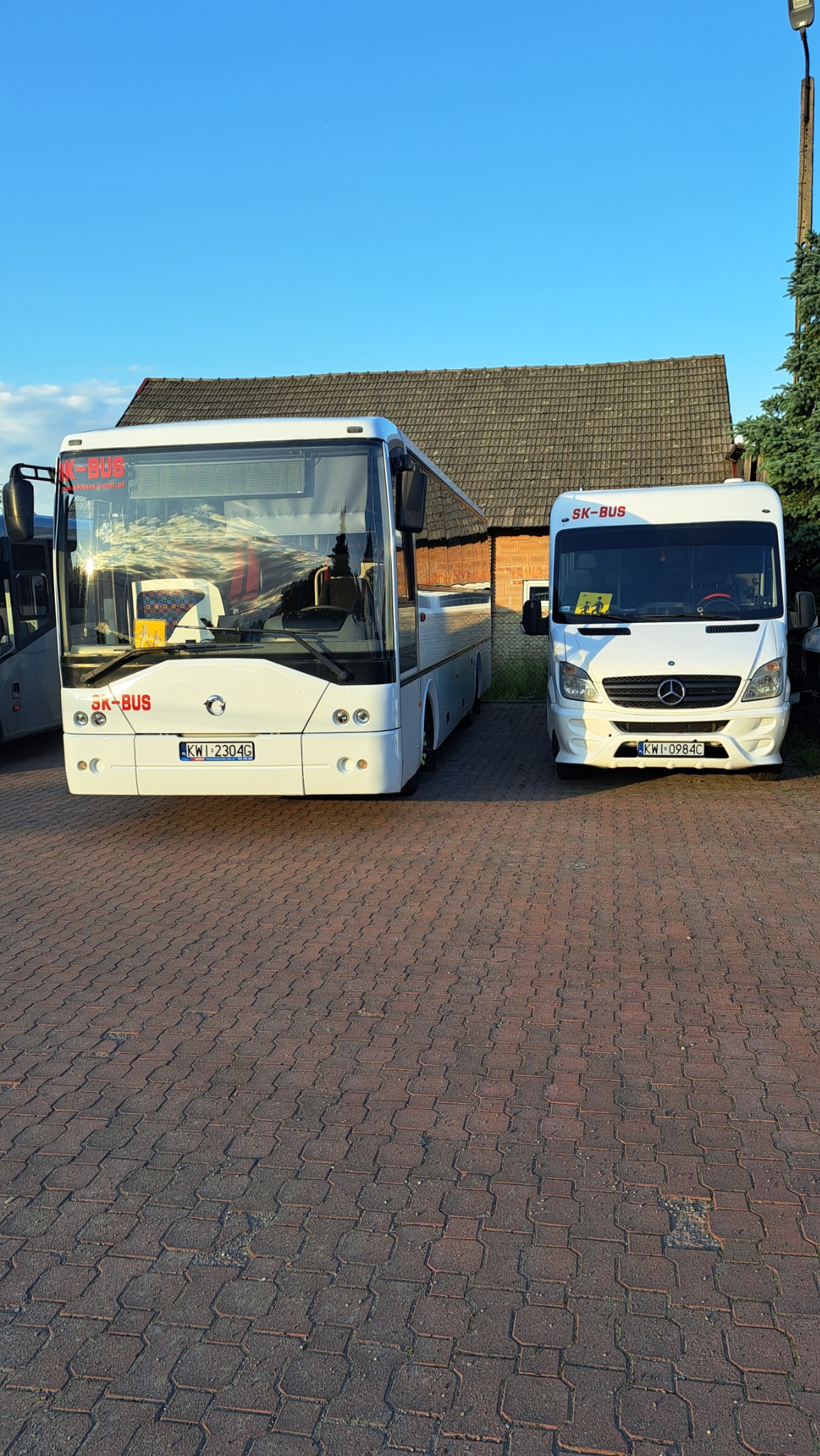 Dwa białe busy z logo SK-BUS zaparkowane na kostce brukowej przed budynkiem z brązowym dachem, widoczne tablice rejestracyjne.