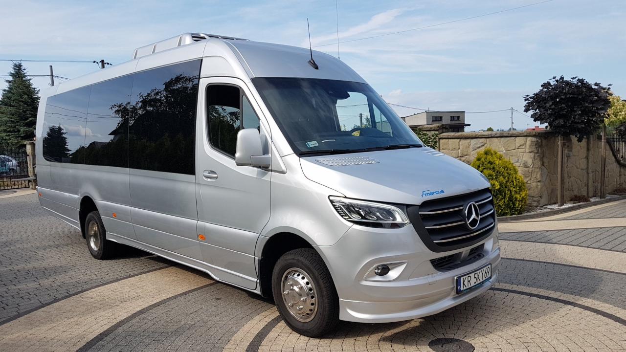 Srebrny bus marki Mercedes Sprinter z przyciemnianymi szybami, zaparkowany na kostce brukowej przed kamiennym ogrodzeniem, z widoczną tablicą rejestracyjną 'KR 5KY69'.