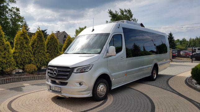 Srebrny Mercedes-Benz Sprinter, luksusowy minibus do przewozu osób, zaparkowany na ozdobnym, okrągłym podjeździe z kostki brukowej.