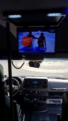 Wnętrze busa z zamontowanym ekranem LCD wyświetlającym reklamę lub film; widoczny kokpit kierowcy i fragment siedzeń.