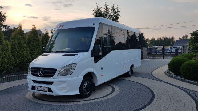 Biały minibus Mercedes-Benz z czarnymi szybami, zaparkowany na ozdobnym brukowanym podjeździe przed budynkiem i zielenią.