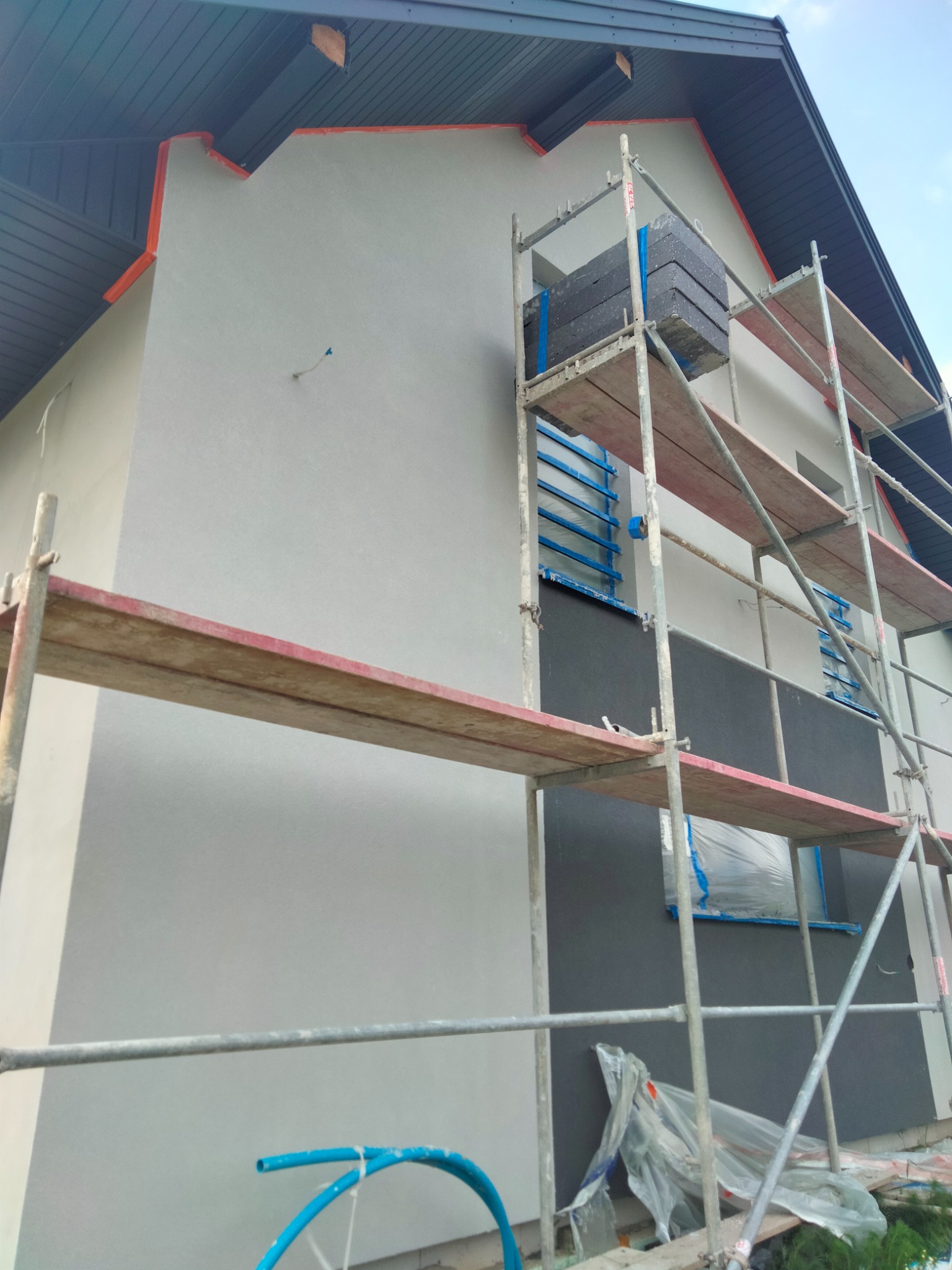 Dom w trakcie ocieplania: część elewacji pokryta szarym tynkiem, część z nałożonym styropianem, rusztowanie z materiałami budowlanymi i zabezpieczonymi oknami.