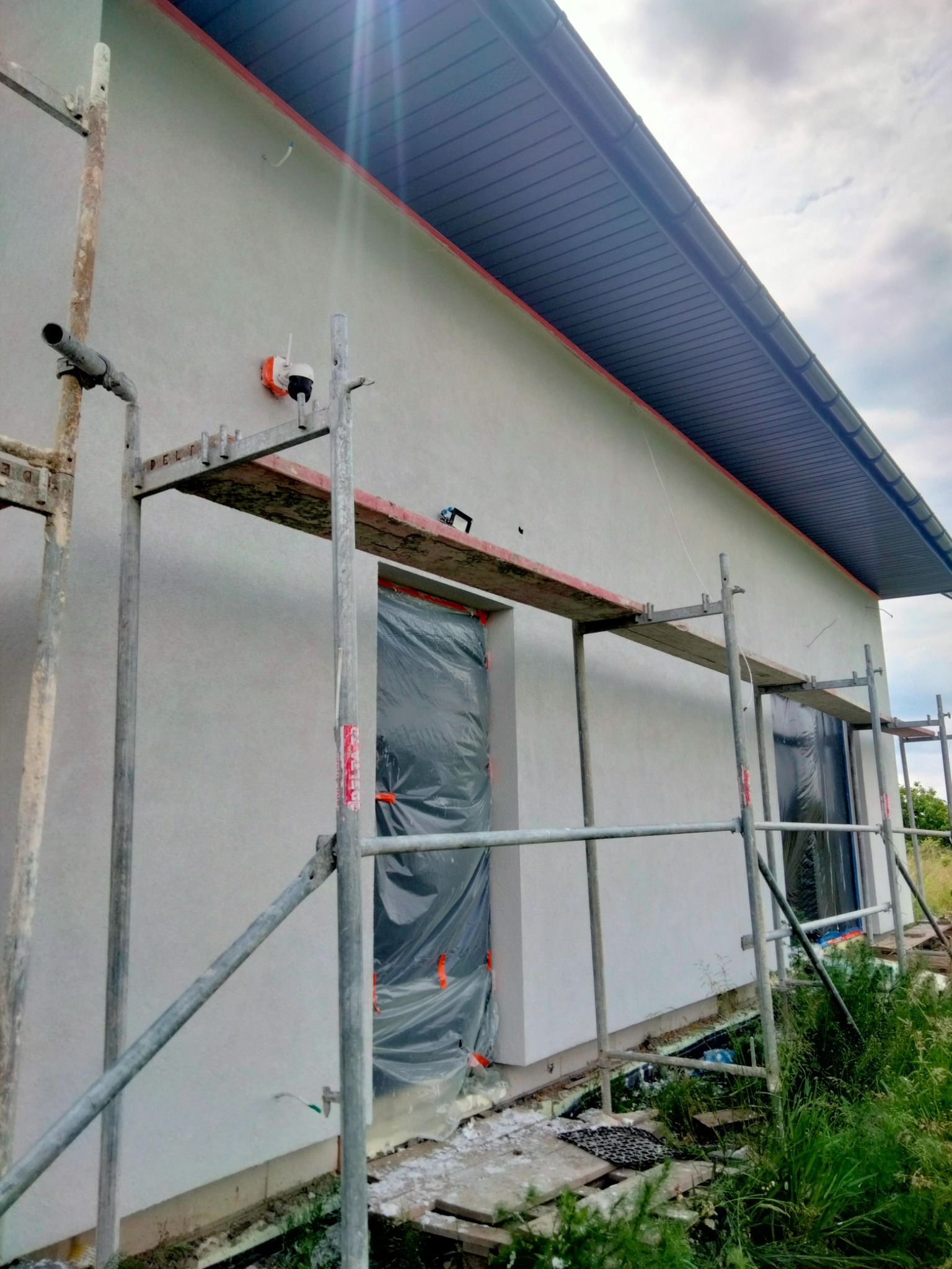 Elewacja domu jednorodzinnego w trakcie malowania, widoczne rusztowanie, okno zabezpieczone folią, świeżo nałożony tynk i wystające kable elektryczne.