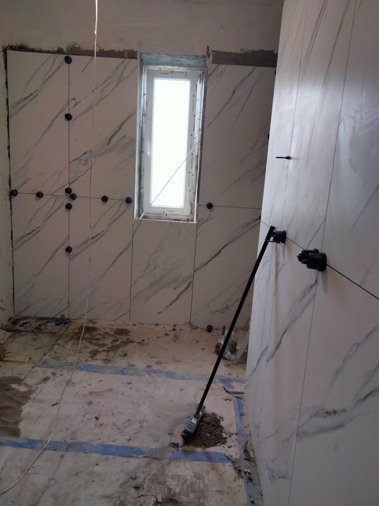 Świeżo ułożone, marmurowe płytki ścienne w trakcie montażu, z użyciem poziomicy laserowej i klipsów dystansowych, widoczne surowe ściany i podłoga w trakcie remontu.