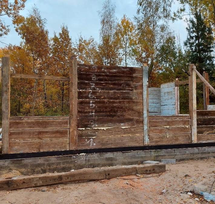 Wznoszenie drewnianej konstrukcji domu, widoczne poziome deski z naniesionymi numerami, fundament z bloczków betonowych, jesienne drzewa w tle.