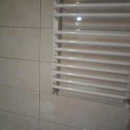 Mariusz Mioduszewski - Biały, drabinkowy grzejnik łazienkowy zamontowany na ścianie z beżowych, poziomych płytek, widoczny z boku fragment drabiny.