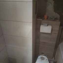 Mariusz Mioduszewski - W trakcie remontu łazienki: widok z góry na niedokończoną toaletę, obok świeżo położone, poziome płytki w jasnym odcieniu, a w tle surowa ściana i narzędzia.