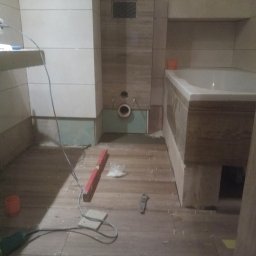 Mariusz Mioduszewski - Łazienka w trakcie remontu: ułożone drewnopodobne płytki podłogowe, poziomica, narzędzia, widoczne przygotowane przyłącza wodno-kanalizacyjne i elektryczne, fragment wanny i zabudowy geberitu.