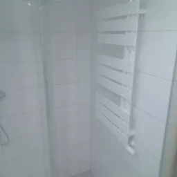 Biały grzejnik drabinkowy zamontowany na kafelkowanej ścianie wewnątrz kabiny prysznicowej z widoczną słuchawką prysznicową.