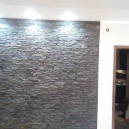 Ściana wyłożona dekoracyjnym kamieniem w odcieniach szarości i brązu, oświetlona od góry punktowymi lampami, z widocznym fragmentem pokoju za otwartymi drzwiami.