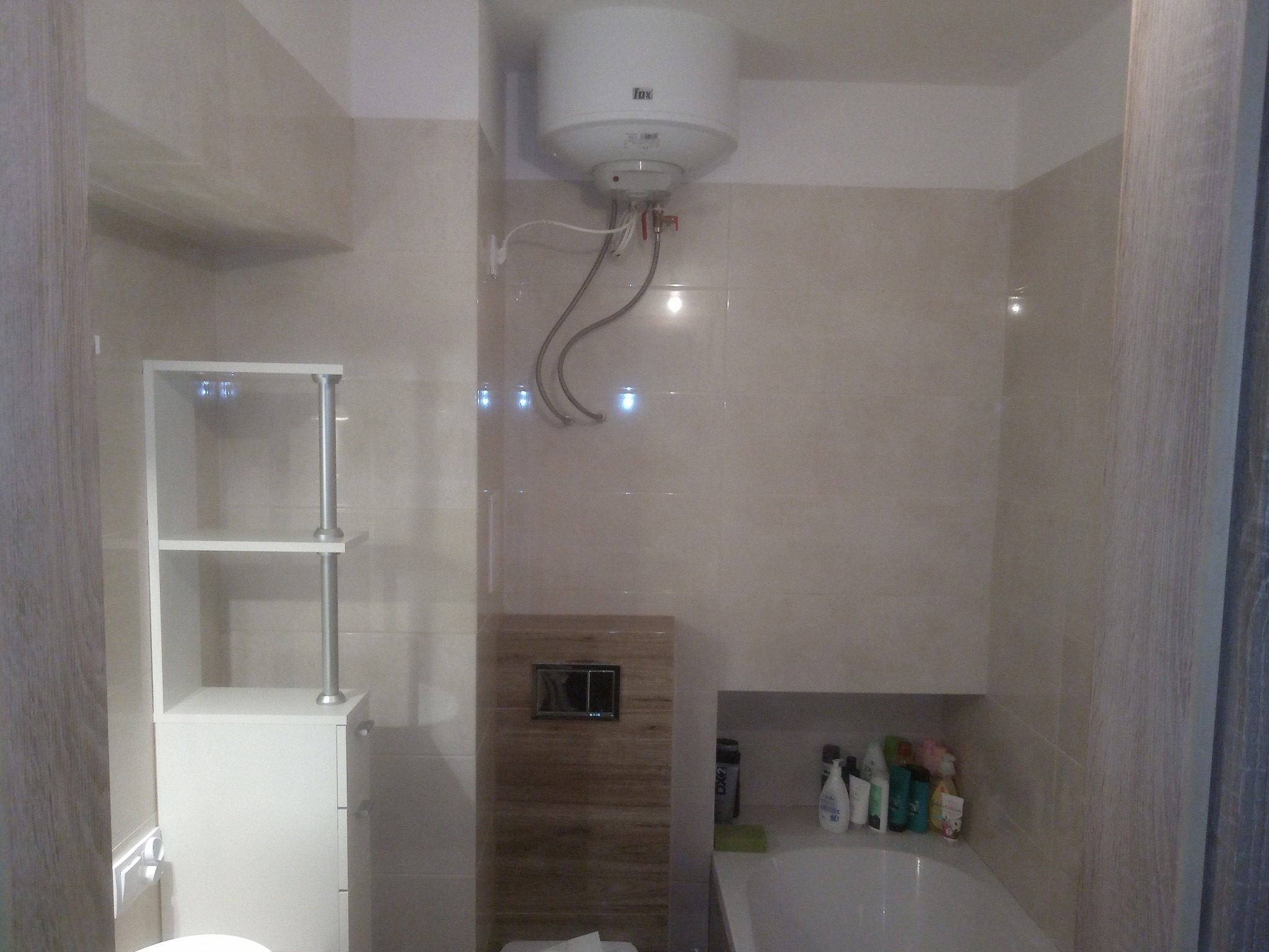 Wykończona łazienka z beżowymi płytkami, zabudowanym geberitem z drewnopodobną okładziną, białym bojlerem elektrycznym i białą szafką z półkami.