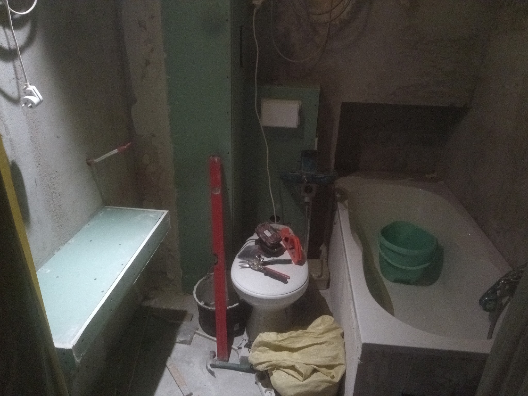 Łazienka w trakcie remontu: widoczne narzędzia na sedesie, poziomica oparta o ścianę, wanna z wiadrem i niedokończona półka z płyt kartonowo-gipsowych.