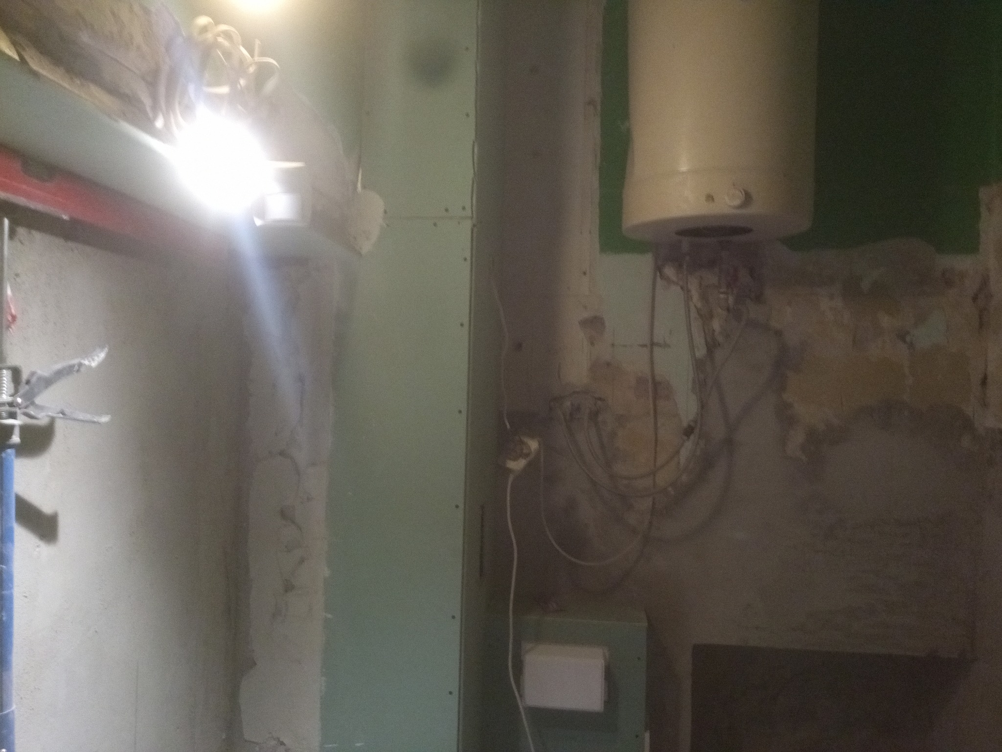 Wnętrze w trakcie remontu: fragment ściany z widocznym bojlerem, wystającymi kablami i fragmentem płyty kartonowo-gipsowej przy słabym oświetleniu.