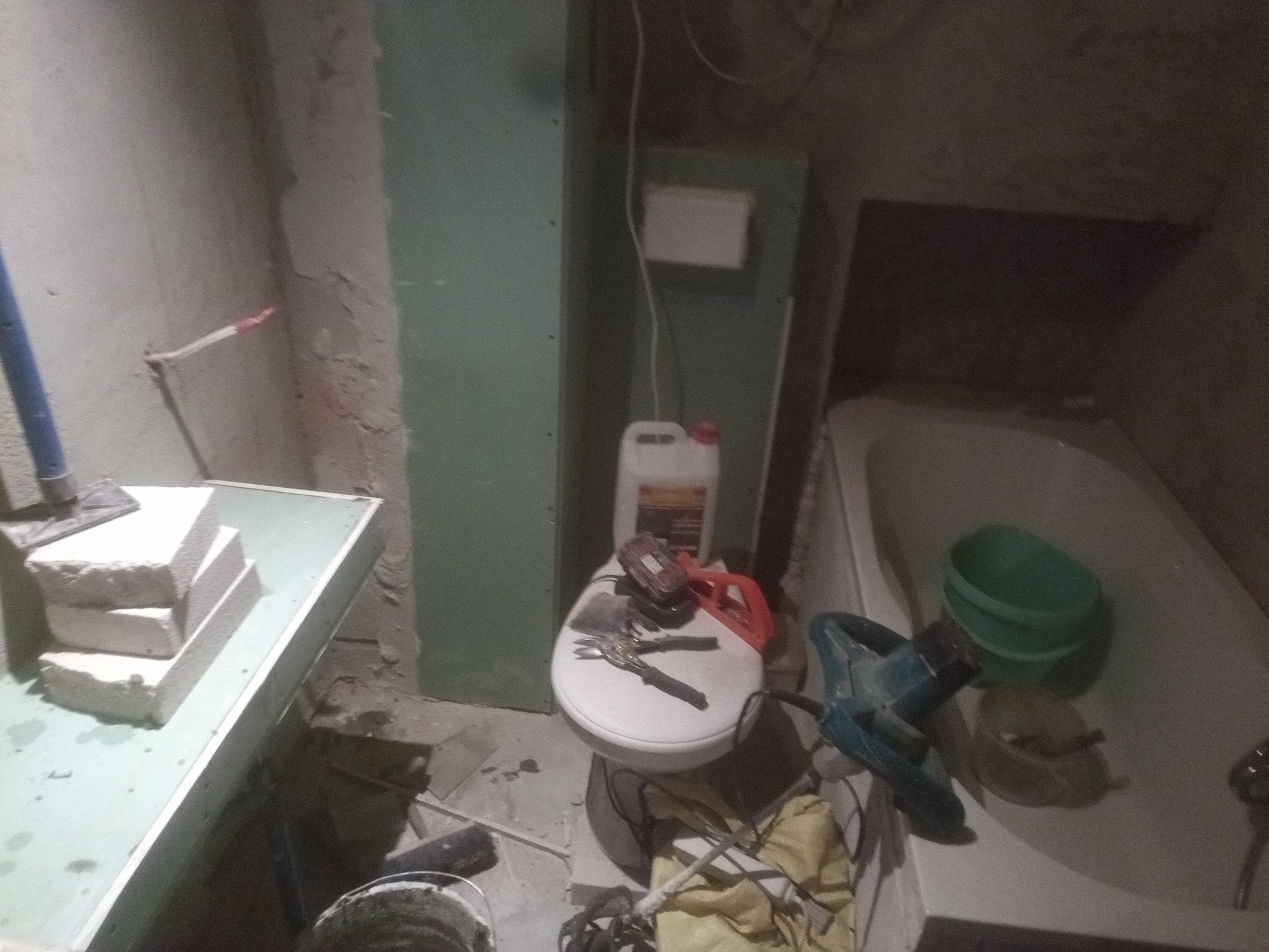 Niedokończony remont łazienki: surowe ściany, płyty gipsowe, narzędzia i materiały budowlane rozłożone na sedesie i w wannie, w tym mieszadło do zaprawy i pojemnik z klejem.