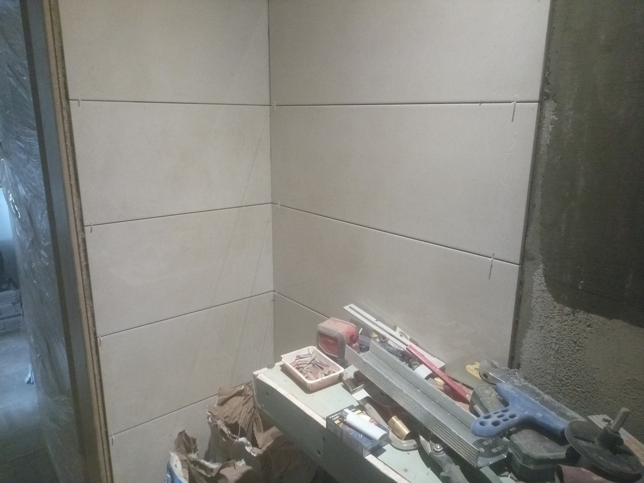 Świeżo ułożone, beżowe płytki na ścianie z widocznymi krzyżykami dystansowymi, narzędzia i resztki materiałów budowlanych na stole roboczym w pomieszczeniu w trakcie remontu.