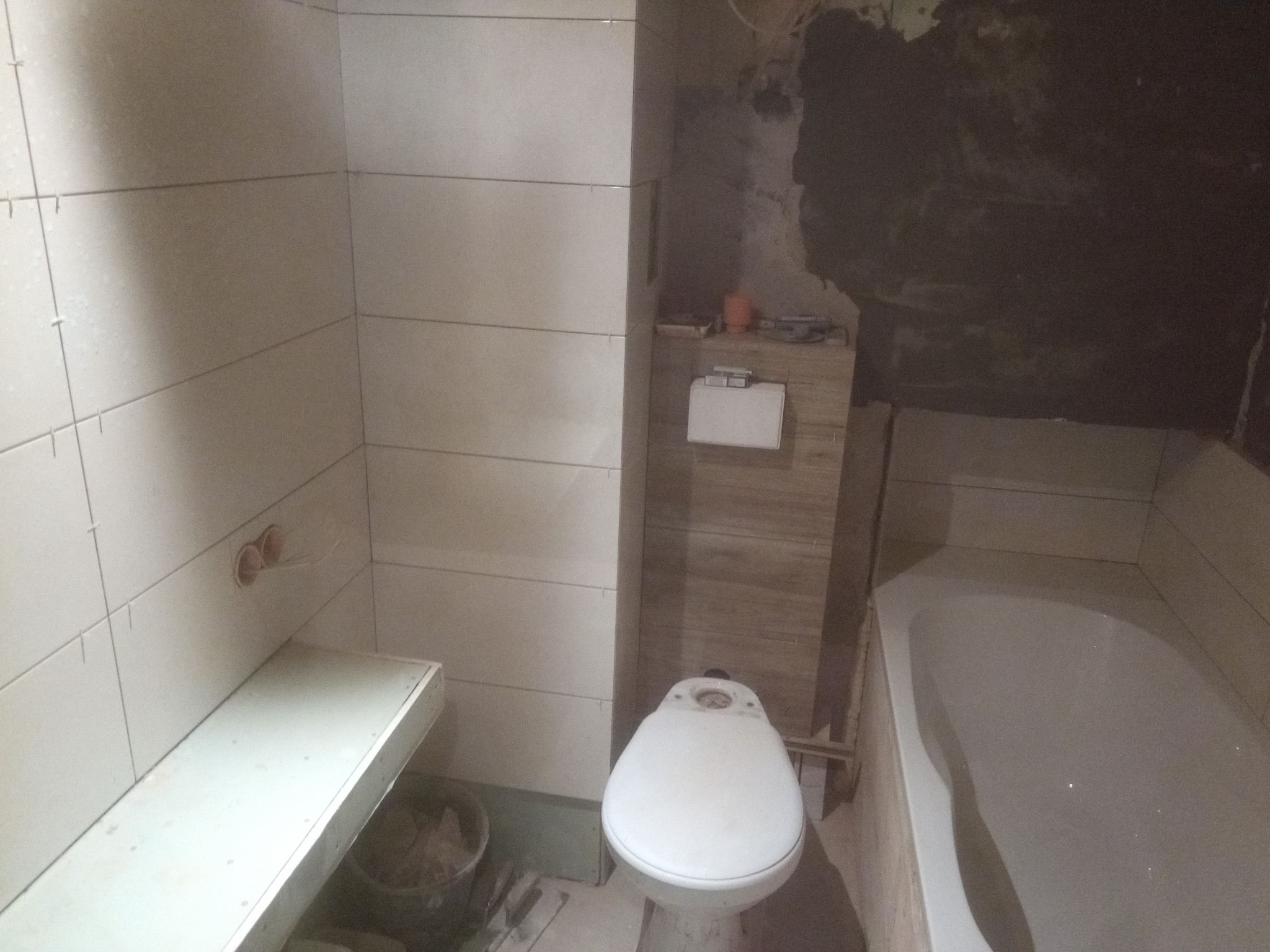 Wnętrze łazienki w trakcie remontu: białe płytki na ścianach, zamontowana toaleta, wanna oraz fragment ściany z surowym tynkiem.