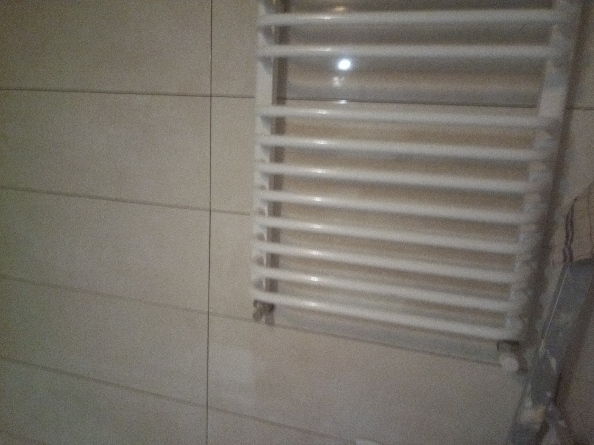 Biały, drabinkowy grzejnik łazienkowy zamontowany na ścianie z beżowych, poziomych płytek, widoczny z boku fragment drabiny.